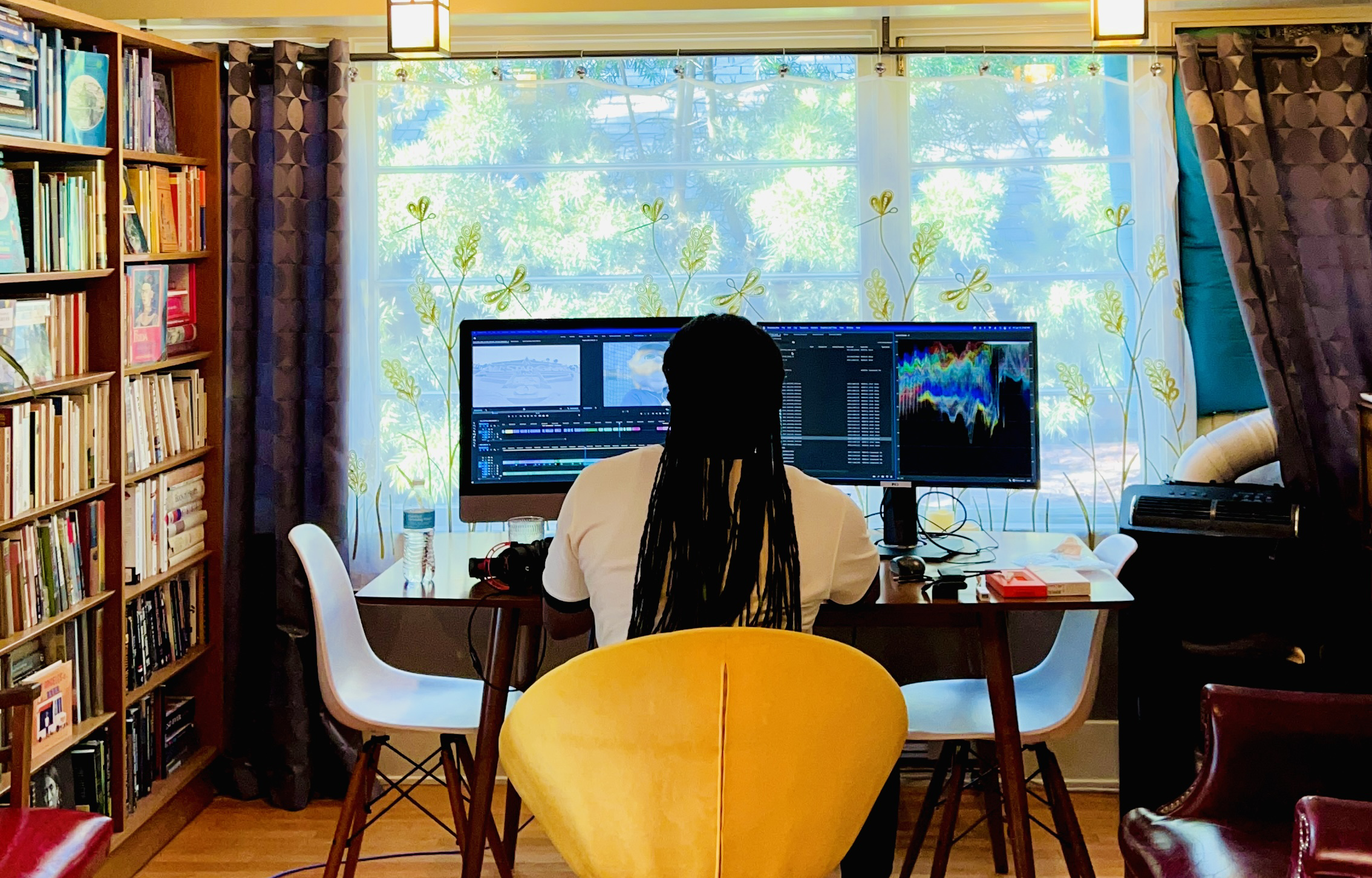 picture of a person editing using two large screens in a room with a view of trees through the window on the other side of the screens
