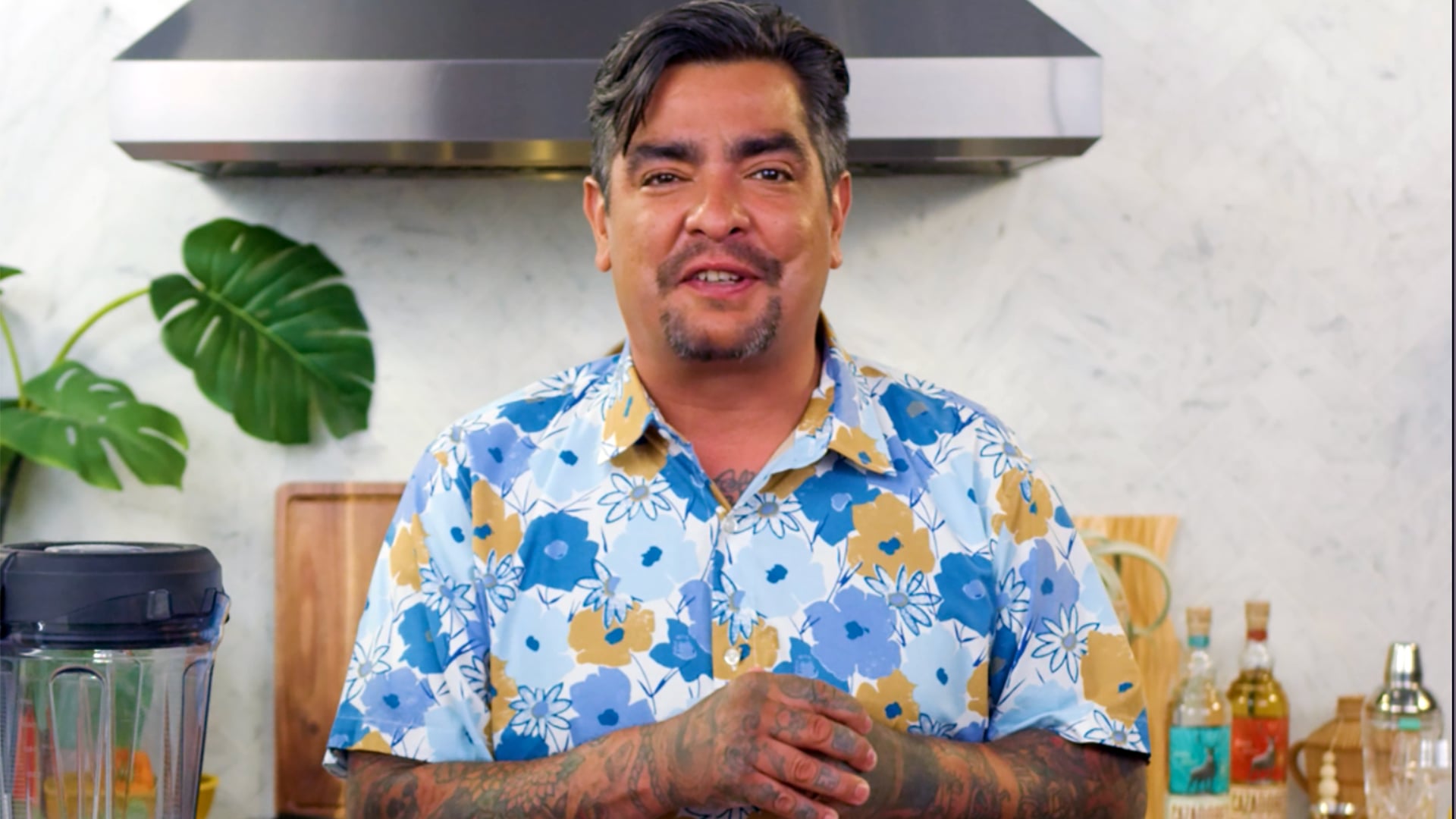 Chef Aaron stands with hands folded on a kitchen set. He wears a brightly colored floral shirt