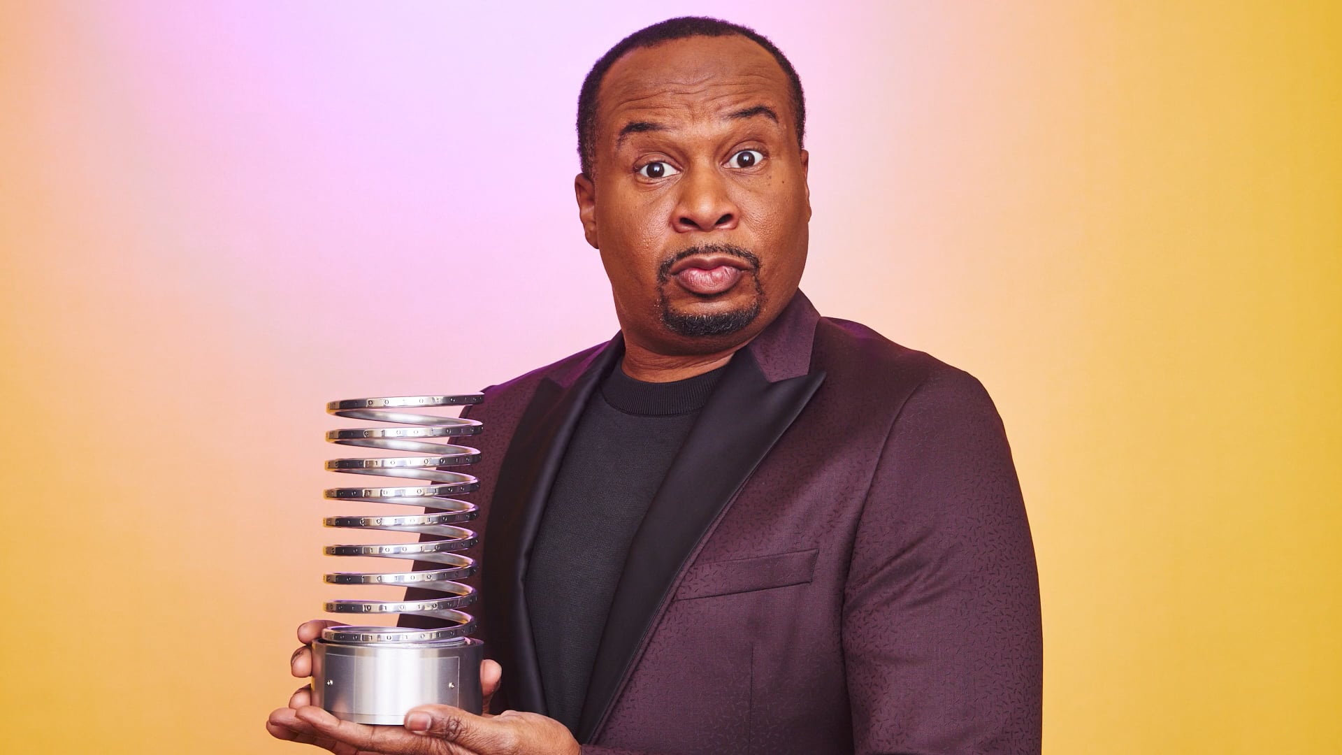 Comedian Roy Wood Jr. wearing a purple tuxedo jacket poses holding a Webby Award in front of a yellow background
