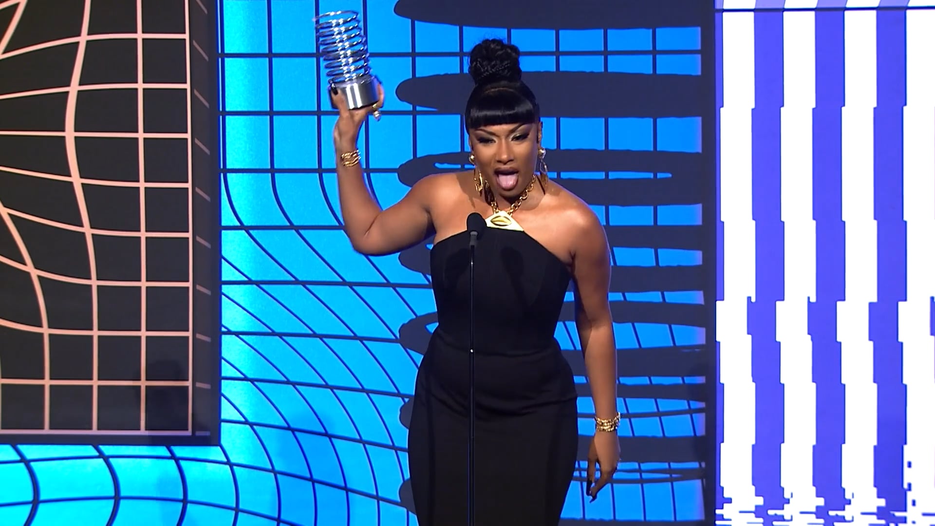 rapper Megan Thee Stallion in a black dress standing on stage in front of a mic holding up her Webby award sticking out her tongue